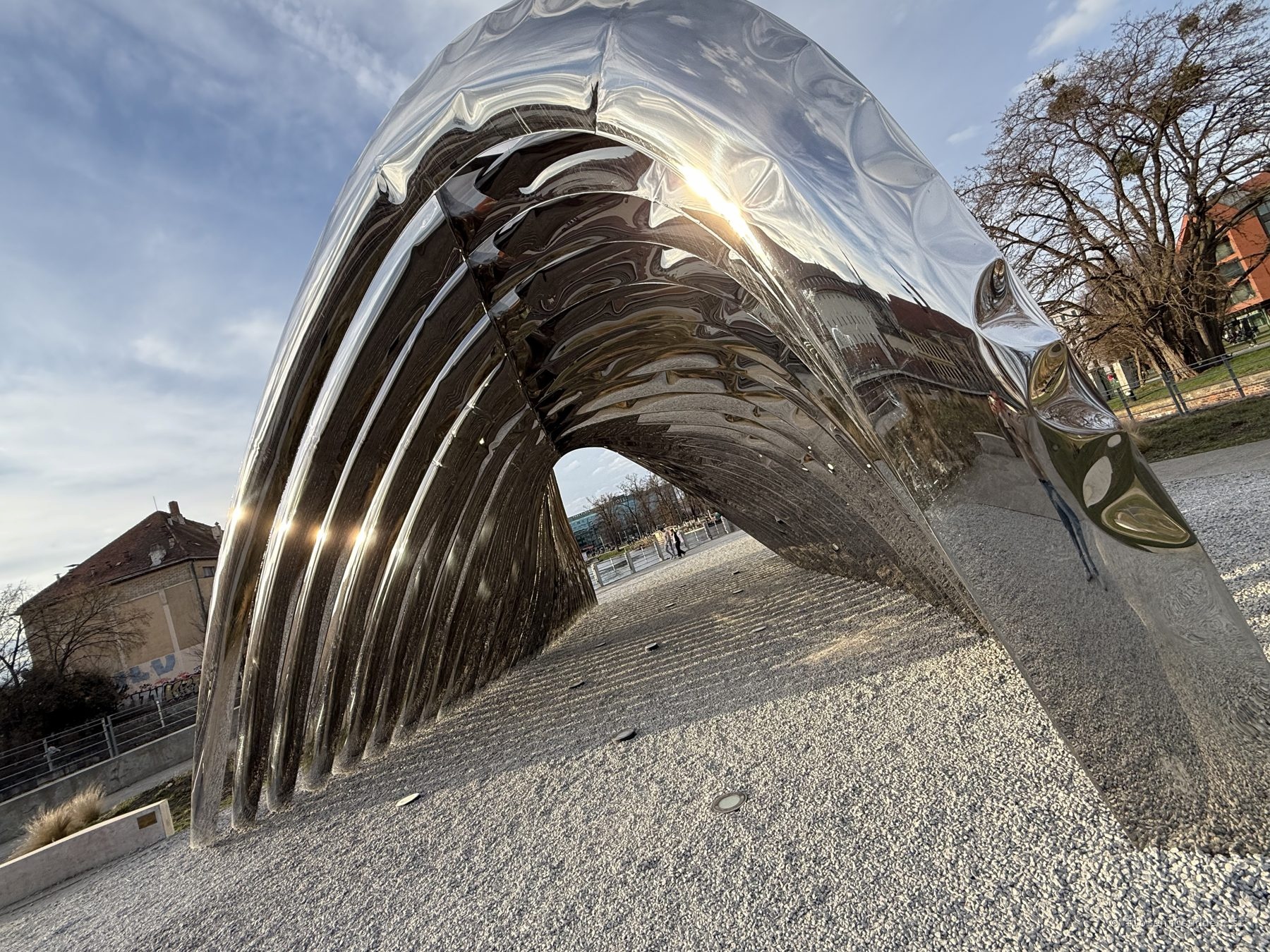 Sculpture contemporaine en acier poli en forme de vague, réfléchissant le ciel et les arbres